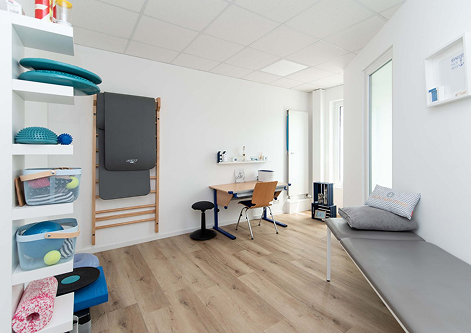 Bright physical therapy room with wooden floor, therapy equipment on shelves and wall, a desk with chair, and a cushioned bench with pillows.