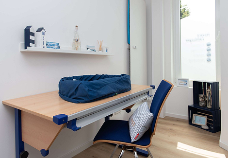Minimalist home office corner with a wooden desk, navy blue chair, pet bed on the desk, and decorative items on a wall shelf and small black cabinet.