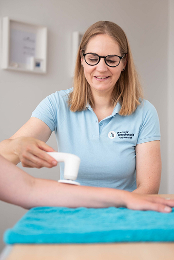 Therapist in glasses using a handheld device on a patient's forearm over a blue towel.