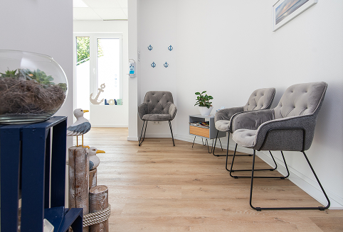 Bright waiting room with three gray cushioned chairs, a small wooden table, nautical decor, and light wood flooring.