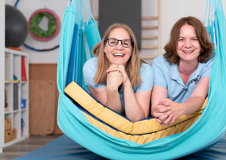 Two smiling women lying on a blue hammock with yellow cushions in a light-filled room with gym equipment.