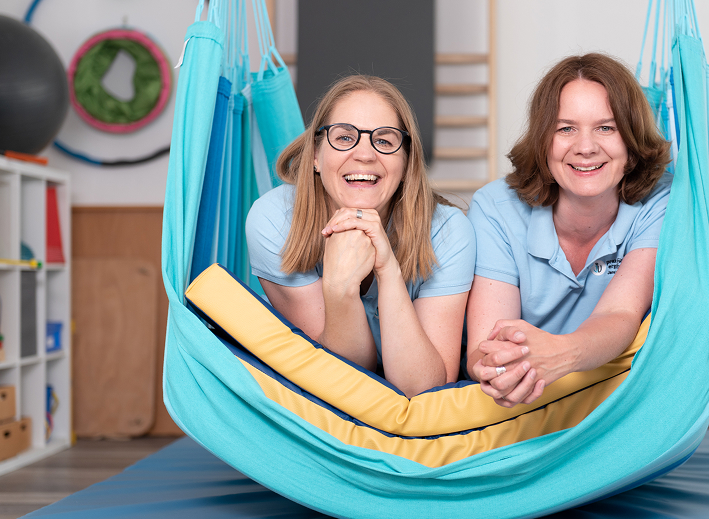 Two women lying side by side in a blue hammock indoors, smiling at the camera.