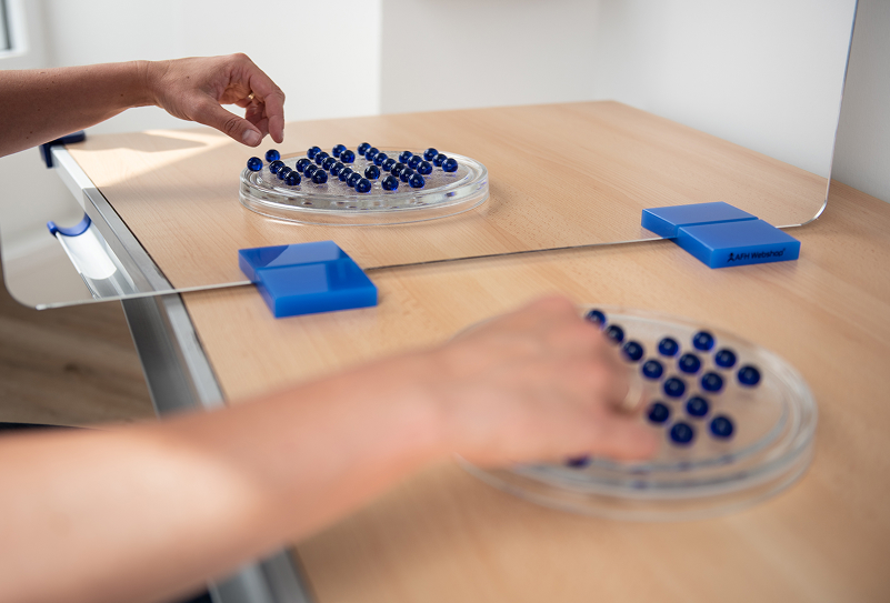 Person placing blue marbles on a clear plastic tray across a transparent divider on a wooden table.