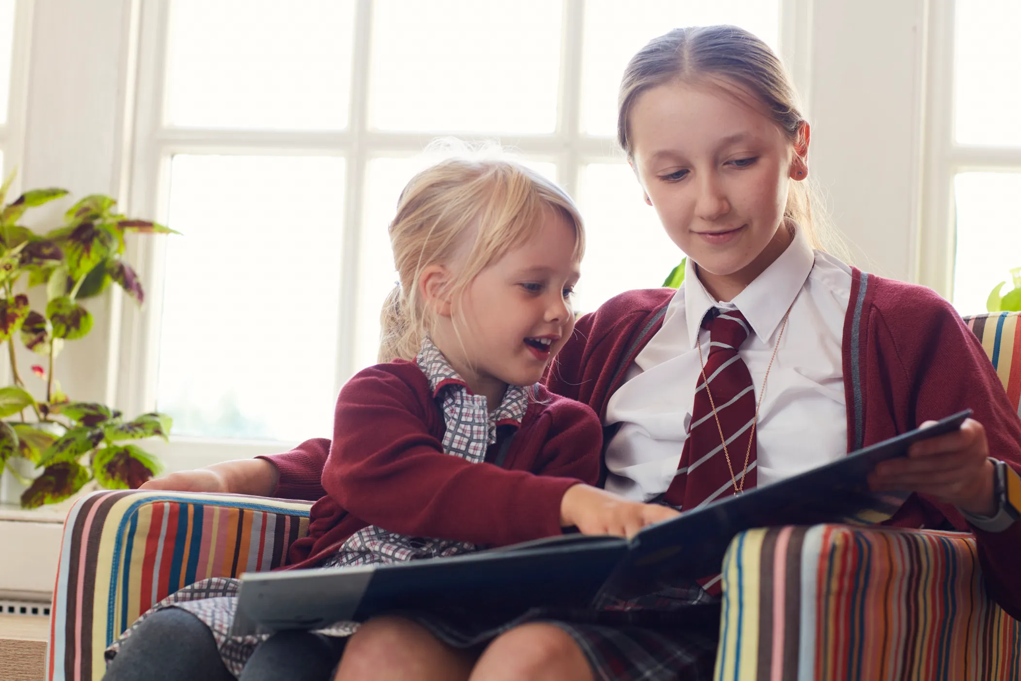 Two girls in school uniforms sitting on a striped armchair, one reading a book to the other.