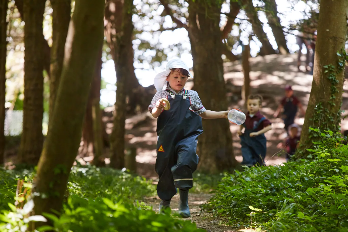 Child wearing waterproof overalls and a sun hat running along a forest path holding a jar and a container.