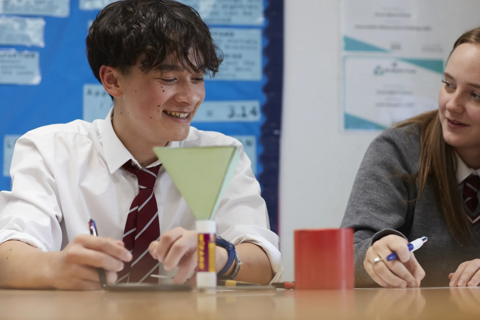 Two students in school uniforms working together at a desk with a glue stick and a red container.