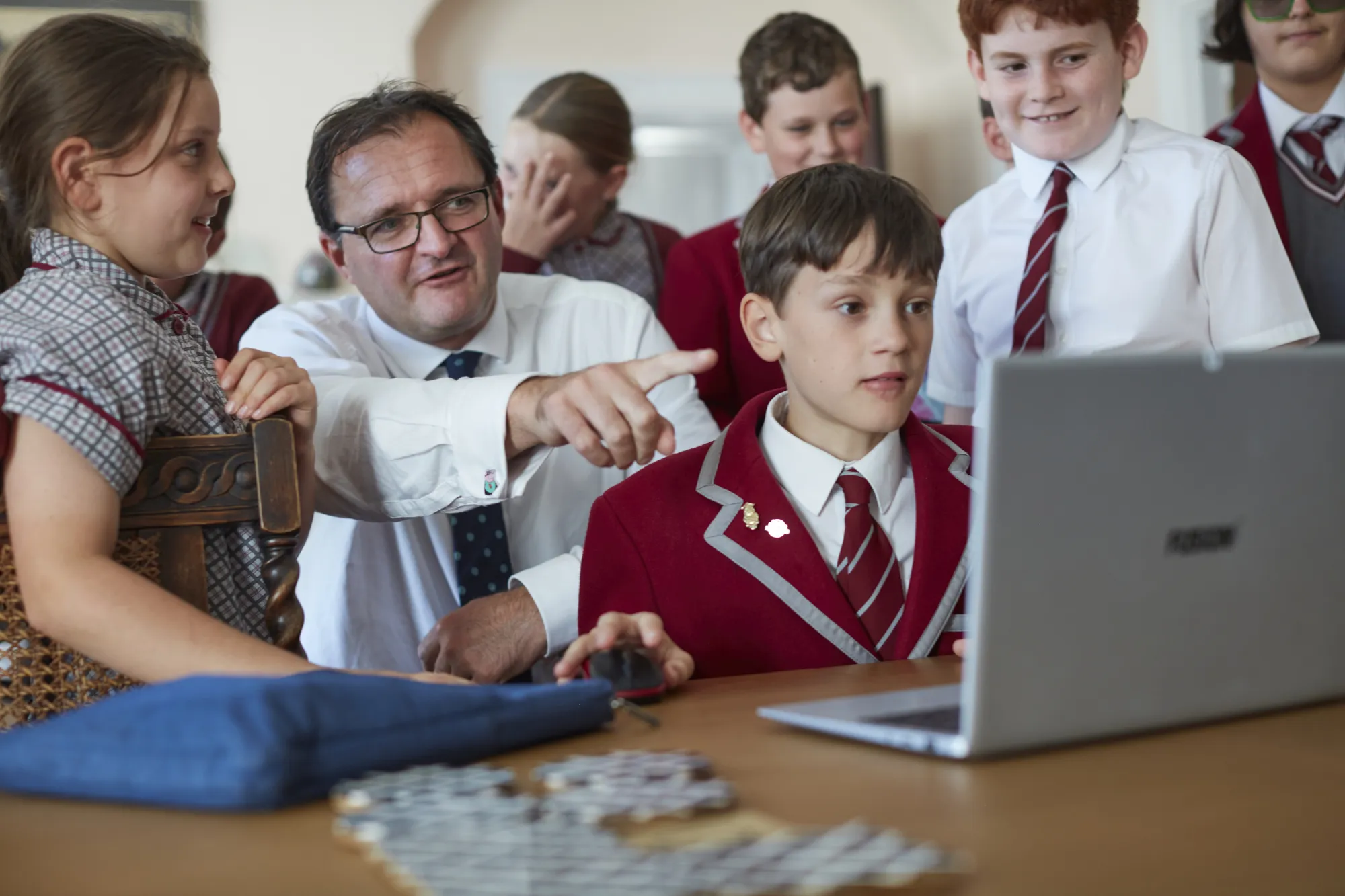 Teacher in white shirt and tie pointing at laptop screen while students in school uniforms watch and smile.