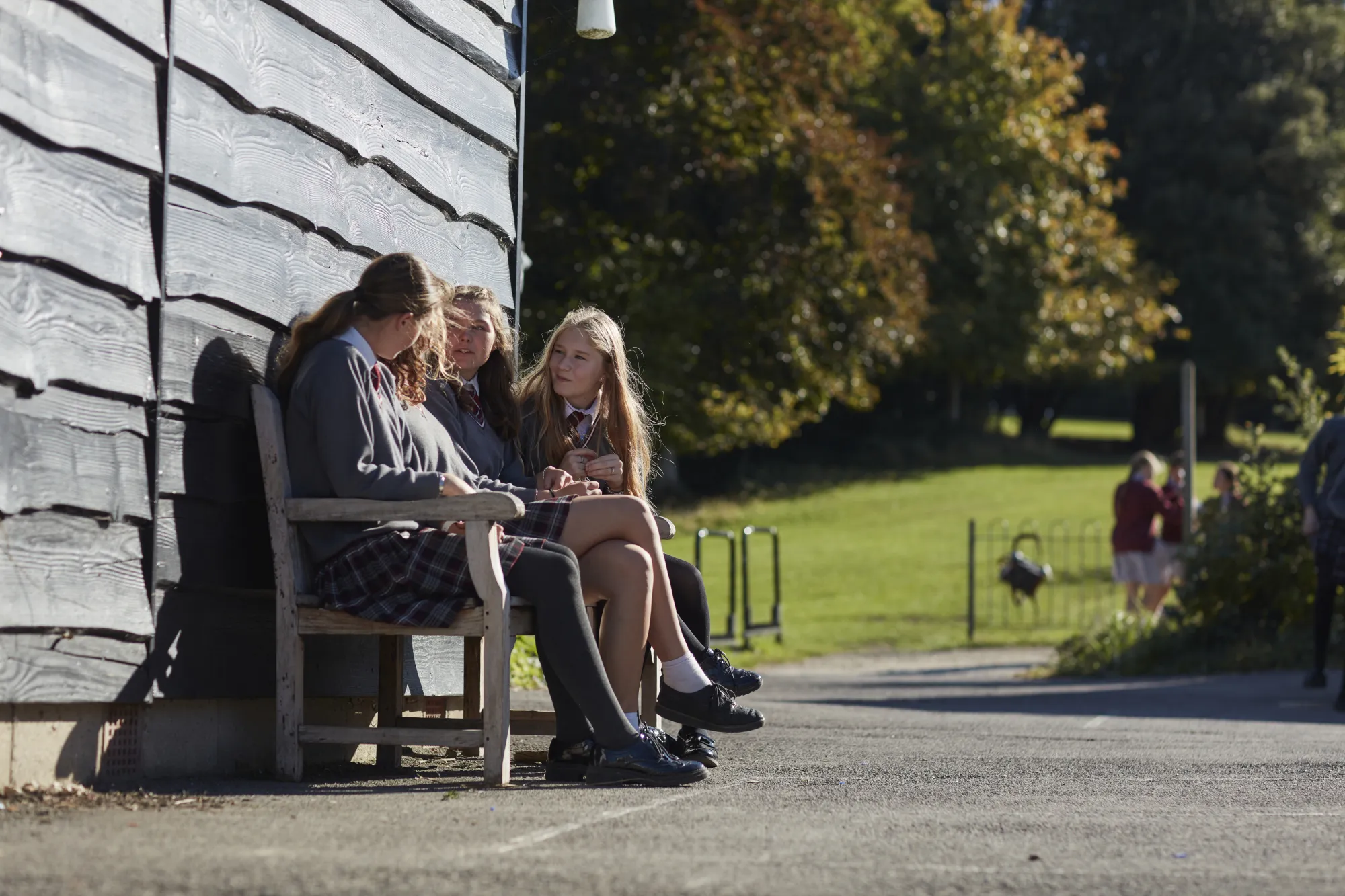 Three schoolgirls in uniform sitting on a wooden bench outside a building, chatting on a sunny day.