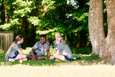 Four children sitting on grass in a circle under trees, enjoying a sunny day outdoors.