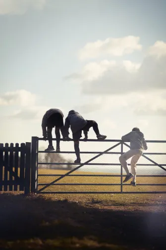 Three people climbing over a metal farm gate into a sunlit field under a cloudy sky.