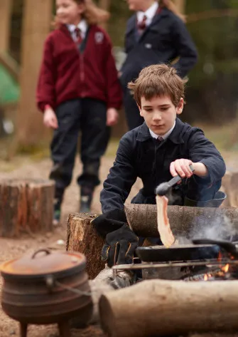 Young boy in school uniform cooking bacon over an open campfire with two other children in the background.