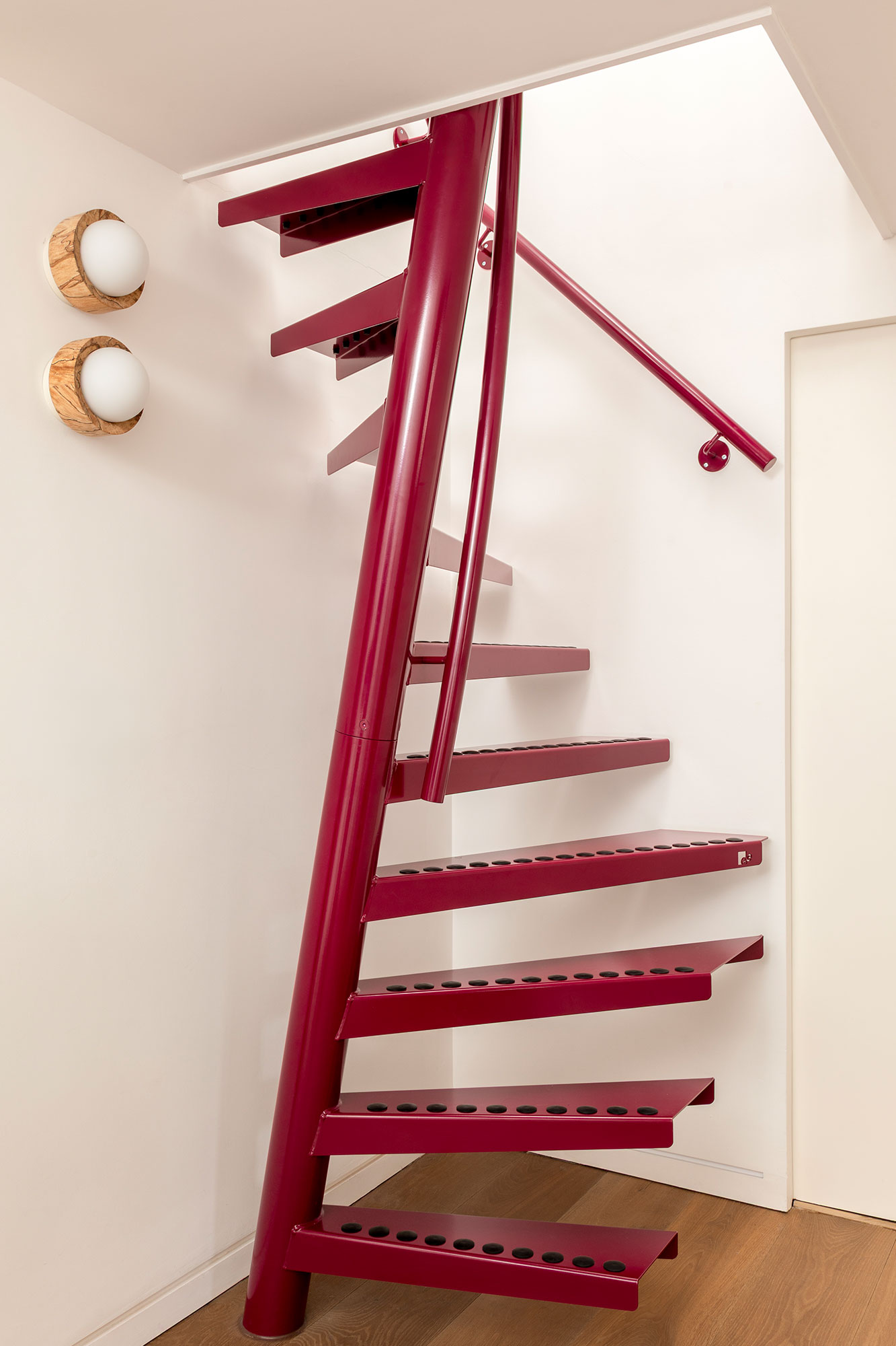 Modern spiral staircase with red metal steps and railing inside a white-walled room with wooden floor.