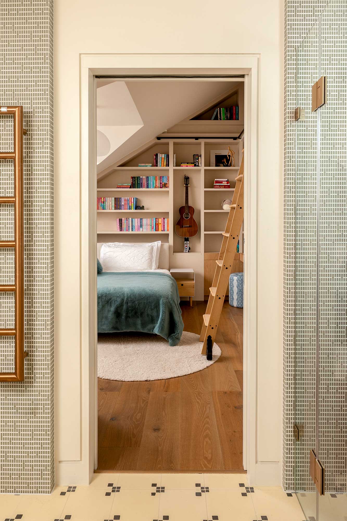 Cozy bedroom with angled ceiling, built-in white shelves filled with books, a wooden acoustic guitar mounted on the wall, a bed with teal bedding and white pillow, a small nightstand, a wooden ladder, and a round beige rug on wooden floor.