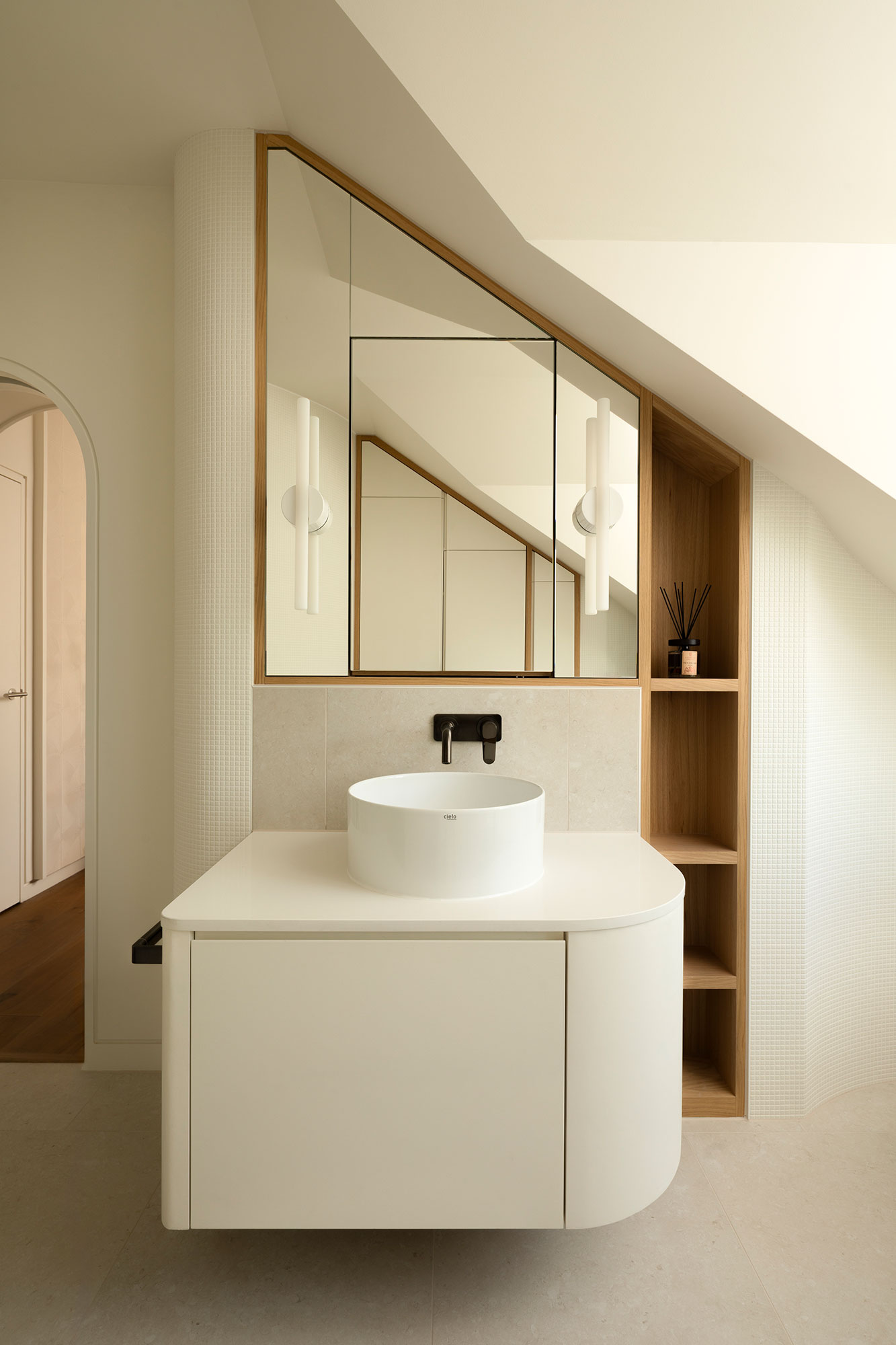 Minimalist bathroom vanity with a round white vessel sink, wall-mounted black faucet, and triangular mirror above it, flanked by wooden shelves.