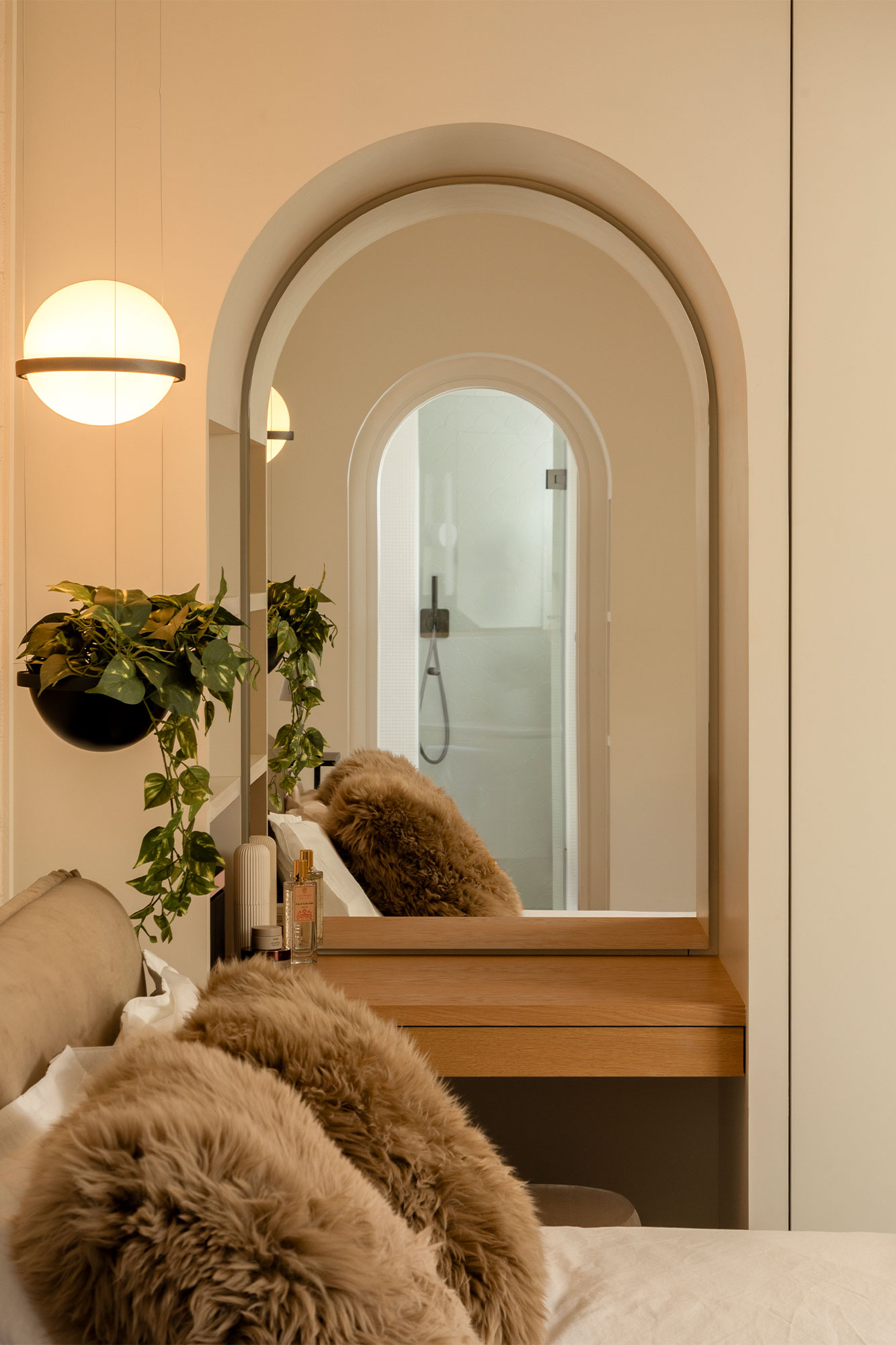 Cozy bedroom corner with a bed featuring fluffy brown pillows, a hanging plant, a round illuminated pendant light, and an arch-shaped mirror reflecting a shower door.