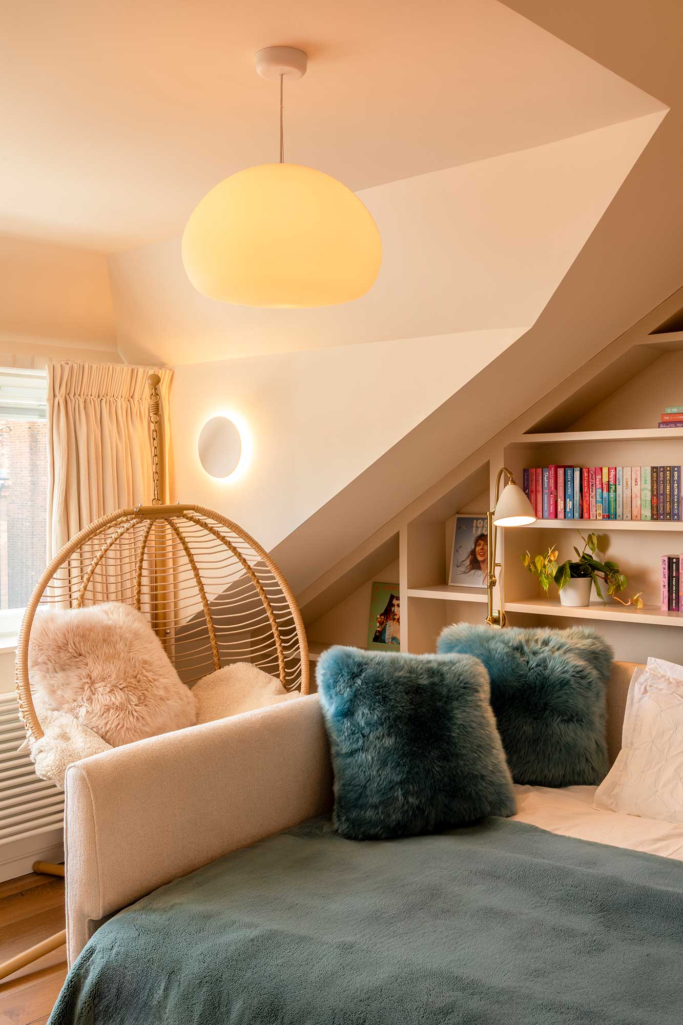 Cozy bedroom corner with a beige sofa bed topped with blue furry pillows, a hanging wicker chair with a fluffy cushion, and shelves with books and plants under a slanted ceiling.