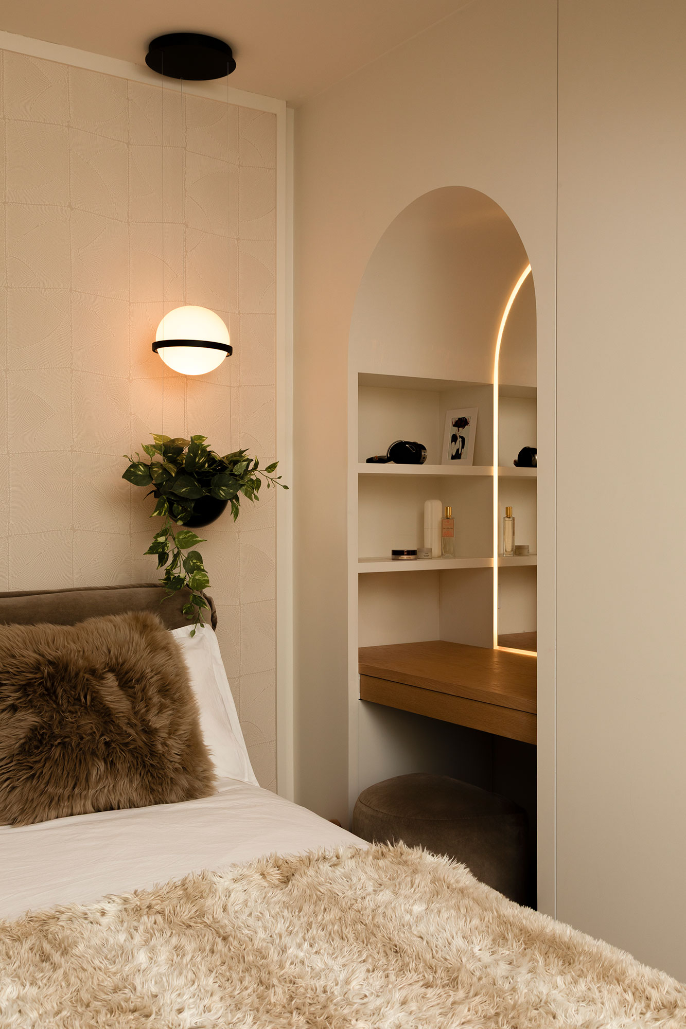 Cozy bedroom corner with beige textured wall, hanging spherical light, trailing green plant, fluffy brown pillow on bed, and an arched built-in vanity with wooden shelf and illuminated mirror.