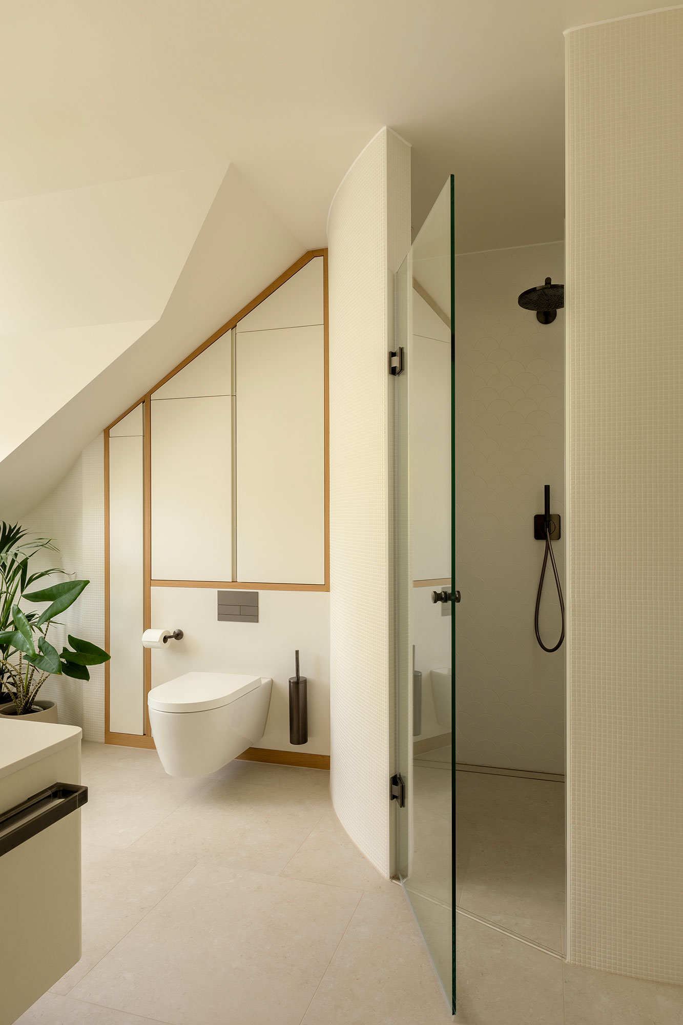 Modern minimalist bathroom with a curved glass shower door, wall-mounted toilet, beige tiles, and a green potted plant.