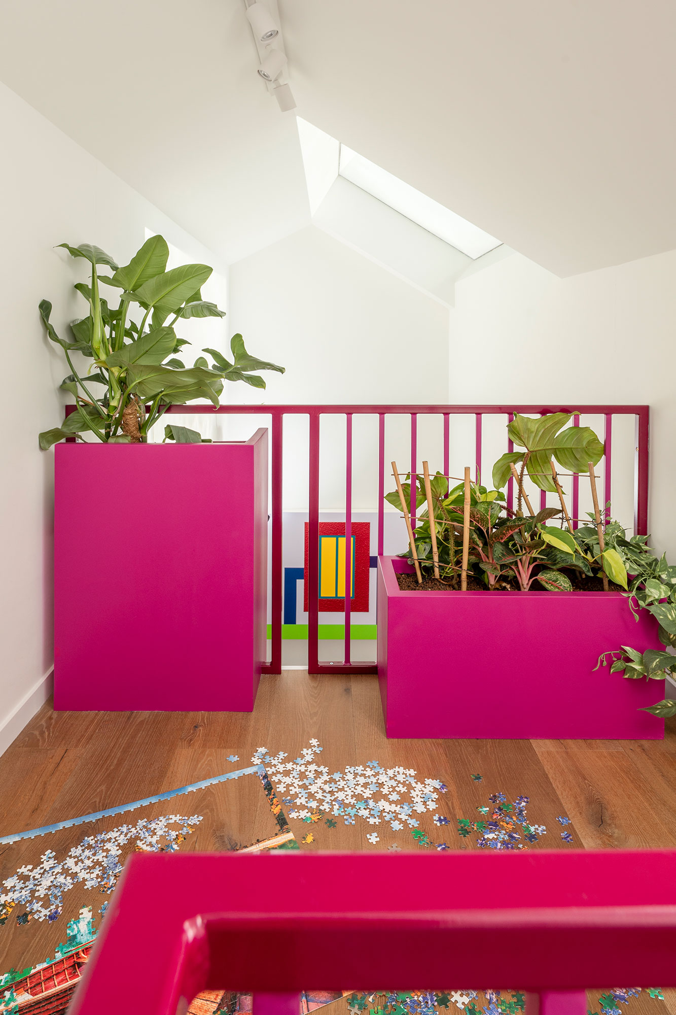 Bright indoor space with wooden floor, two large magenta planters with green plants, a magenta railing, and scattered puzzle pieces on the floor.