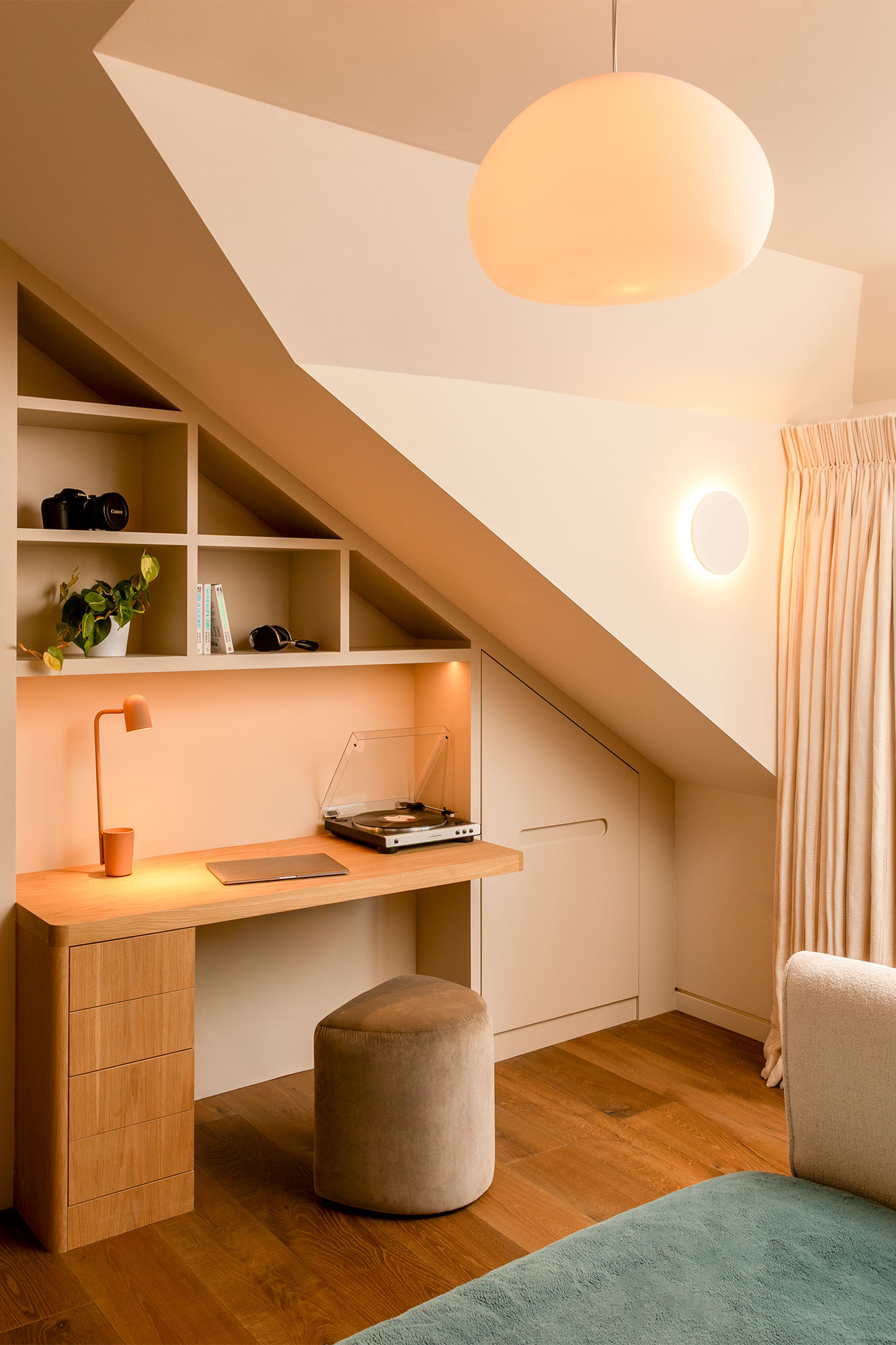 Cozy workspace built under a sloped ceiling with wooden desk, beige stool, record player, lamp, potted plant, and shelving.