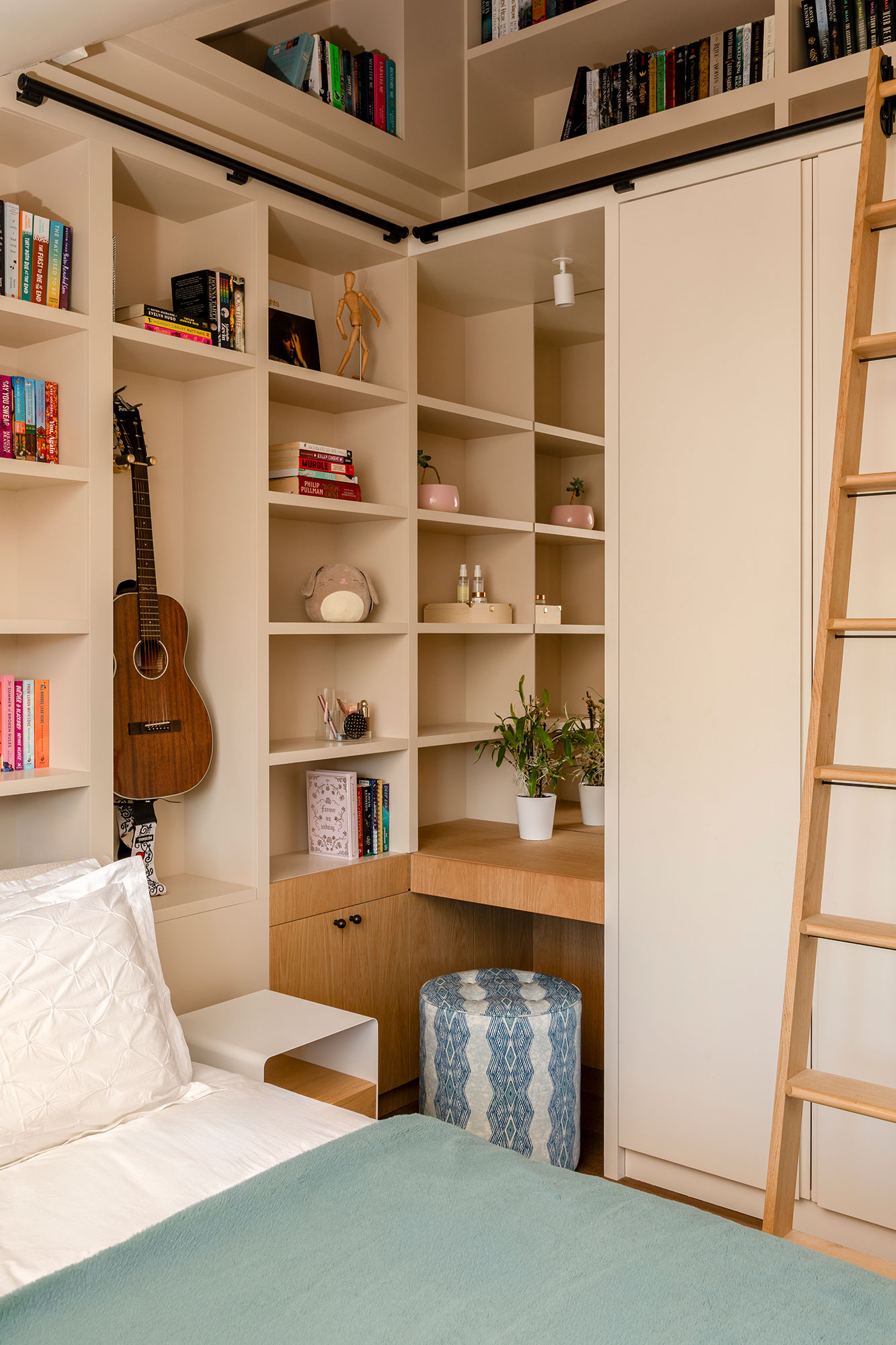 Cozy bedroom corner with built-in beige shelves holding books, a guitar, plants, and decorative items, featuring a blue patterned stool and wooden ladder.