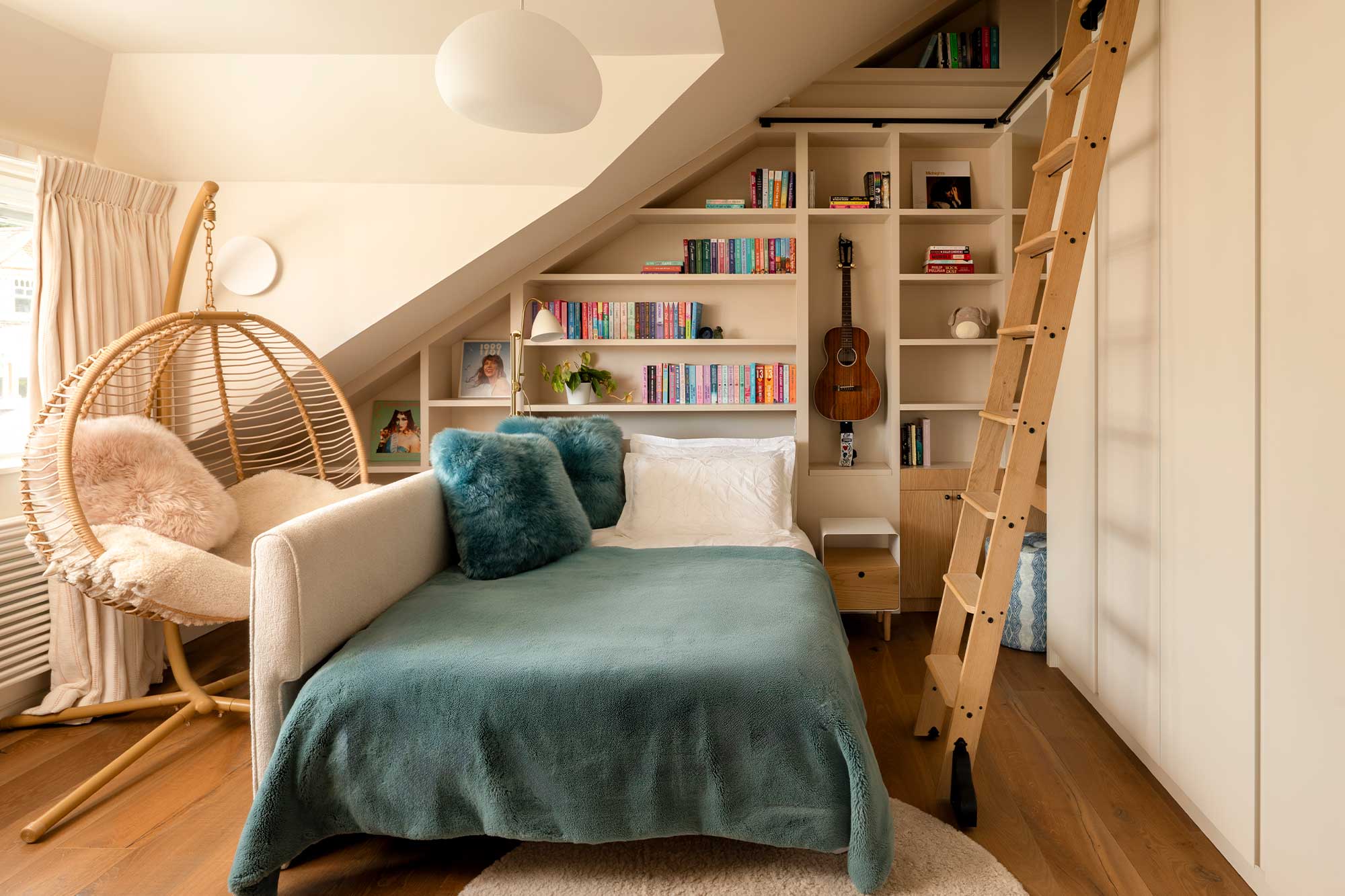 Cozy attic bedroom with a teal blanket, two blue fuzzy pillows, a hanging wicker chair with pink cushions, built-in white shelves filled with books, a guitar, and a wooden ladder.