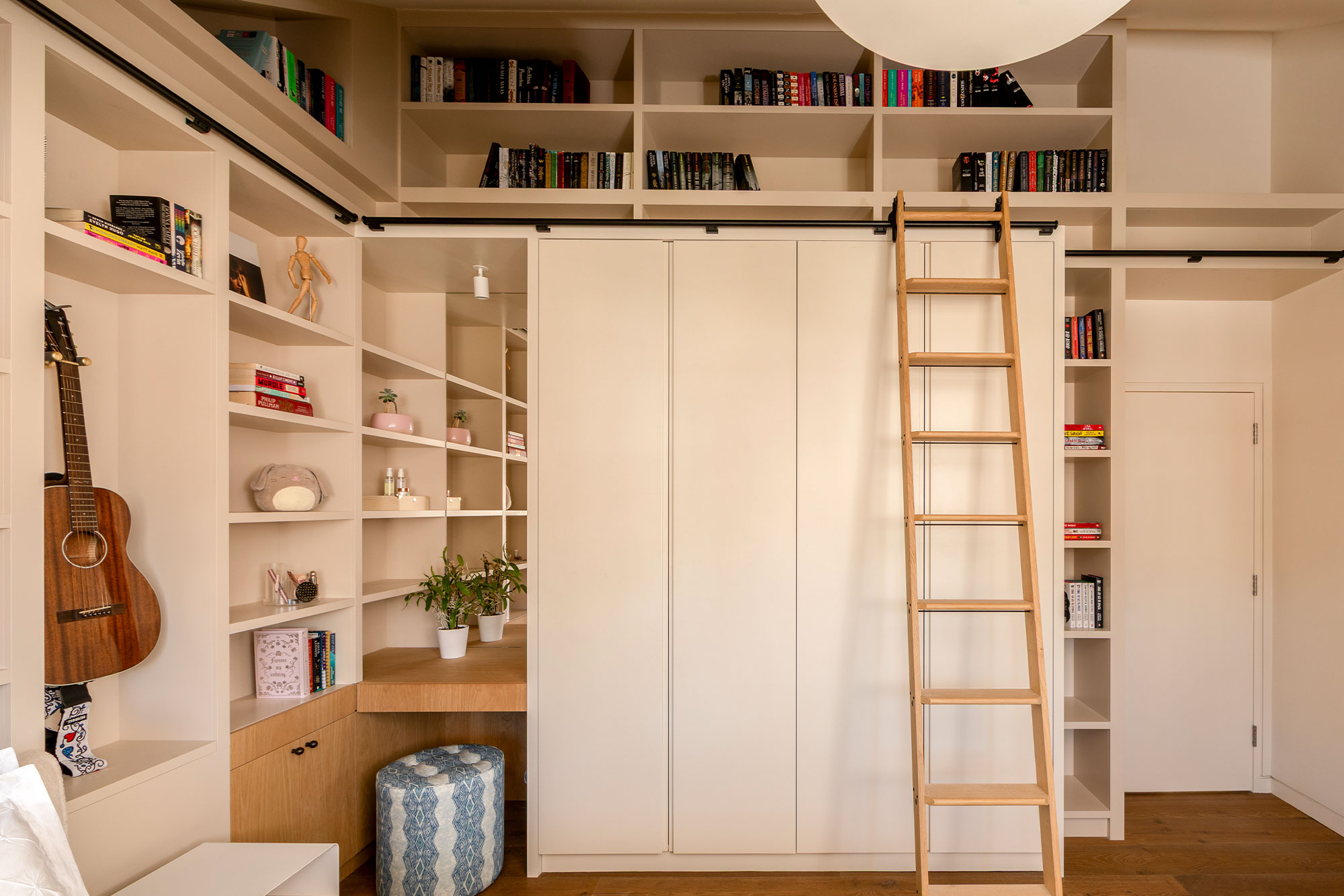 Modern cozy room with built-in beige shelving filled with books, plants, a guitar, a wooden ladder, and a blue patterned ottoman.