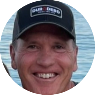 Smiling man wearing a black cap with 'DURO DERO' logo, with water in the background.