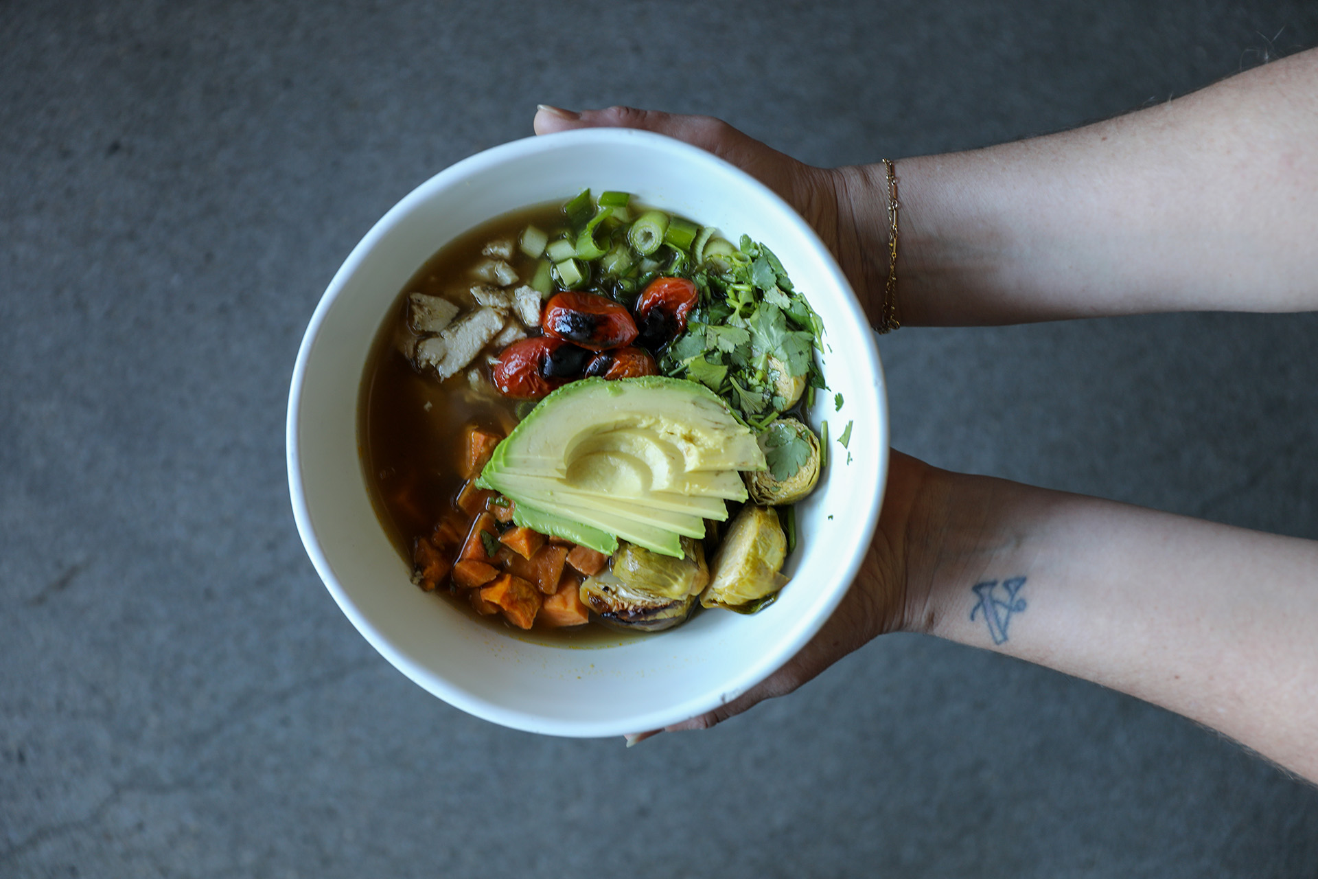 Hands holding a bowl of broth with sliced avocado, grilled tomatoes, Brussels sprouts, sweet potatoes, chopped chicken, green onions, and cilantro.