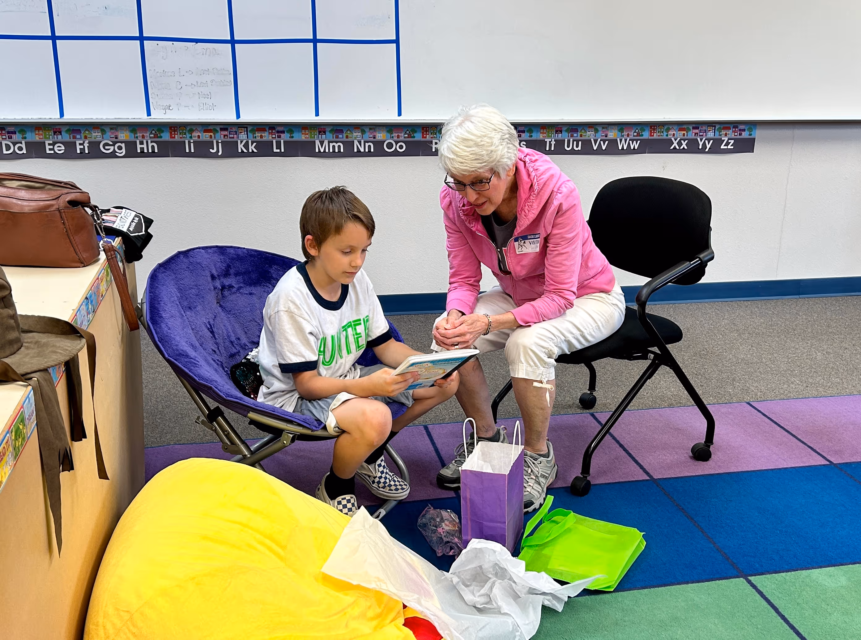 A older lady with short grey hair reading to a little boy in a classroom.