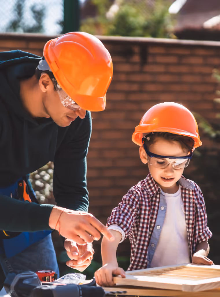 A man with a child, both wearing orange hard hats building something out of wood.