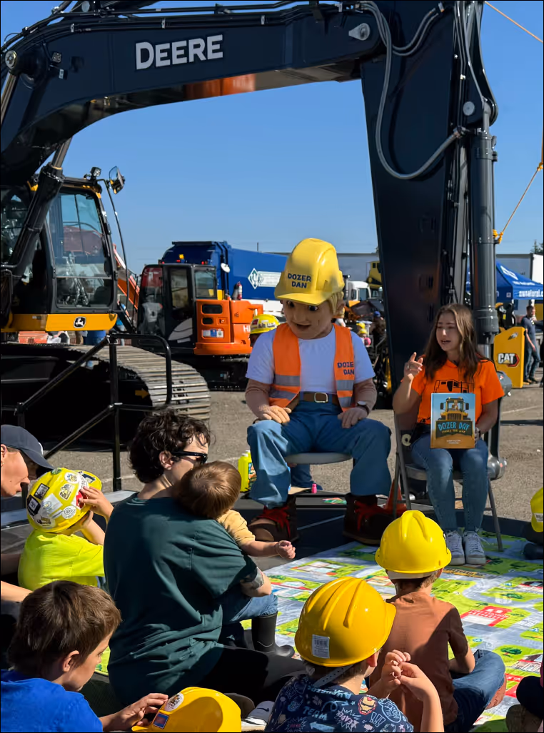 Dozer Dave sitting with a young girl reading a story to children and sitting in front of a large excavator.