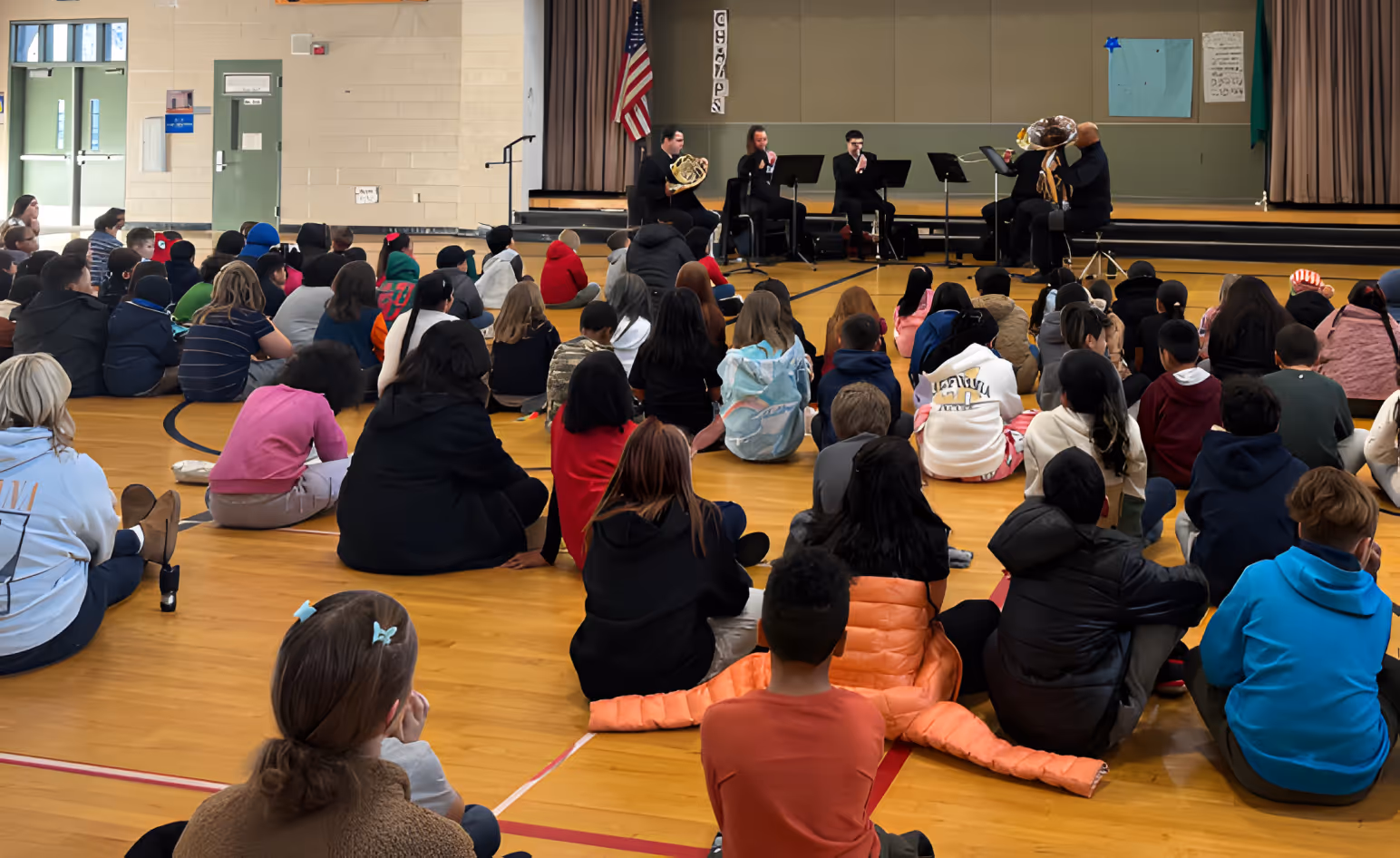 Children seated on the floor of an auditorium, attentively watching a live orchestra performance on stage.