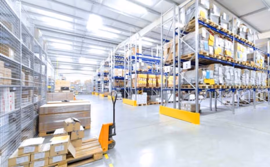 Bright warehouse interior with metal shelving stocked with boxes and a manual pallet jack carrying boxes on a wooden pallet.