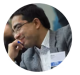 Man wearing glasses and a gray blazer, smiling while resting his chin on his hand.