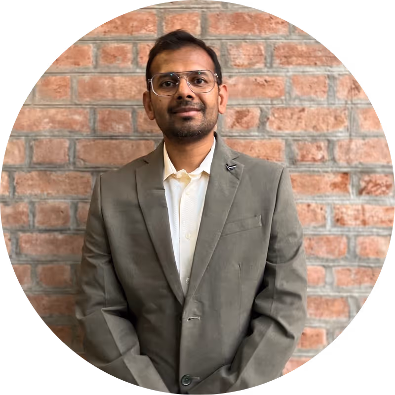 Man wearing glasses and a green blazer standing in front of a brick wall.