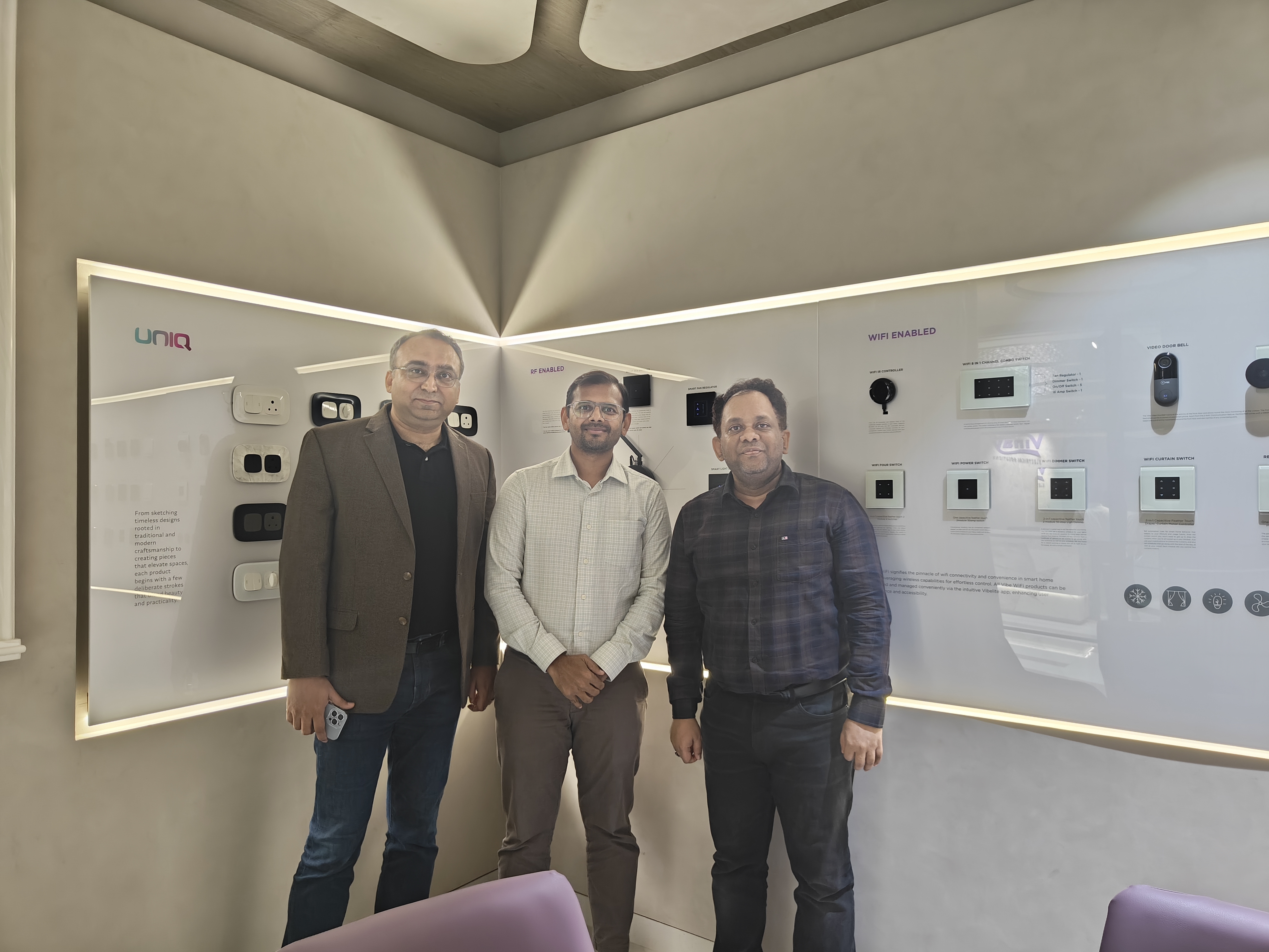 Three men standing in front of a display wall with electrical switches and panels labeled UNIQ and WIFI enabled.