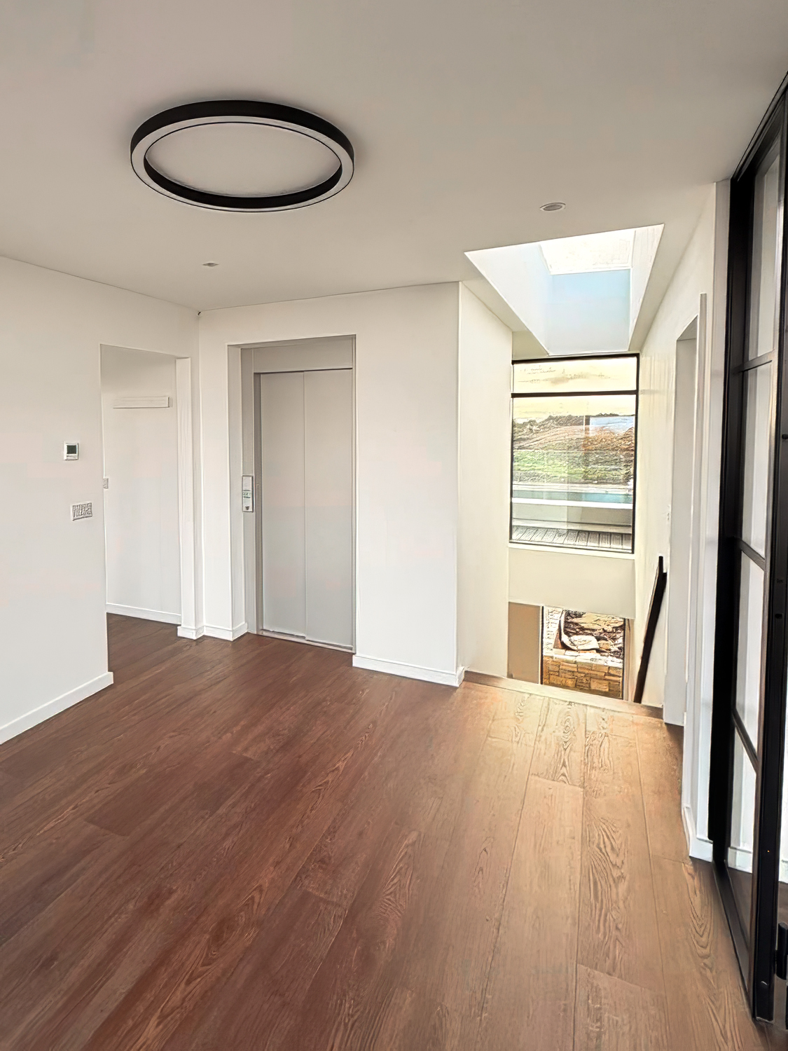 Modern indoor hallway with wooden floors, white walls, an elevator, a skylight, and a large window overlooking an outdoor area.
