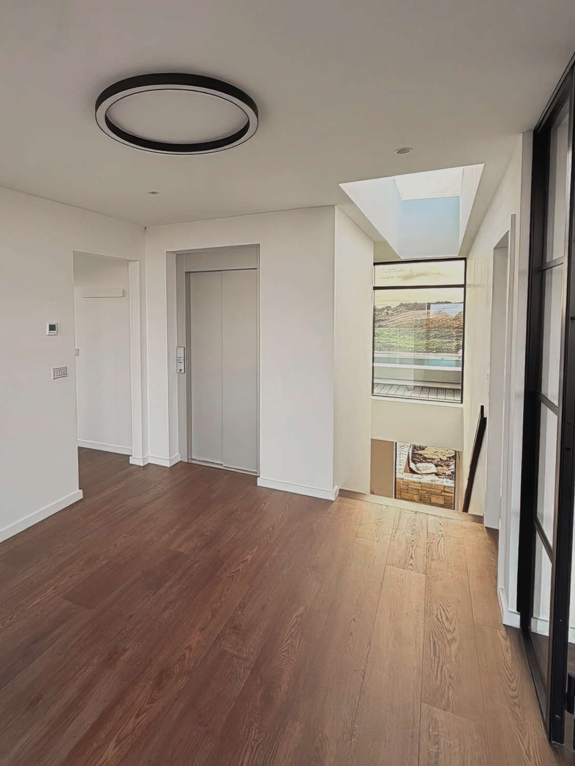 Modern indoor space with wooden flooring, a closed elevator door, a skylight window, and a glass door on the right.