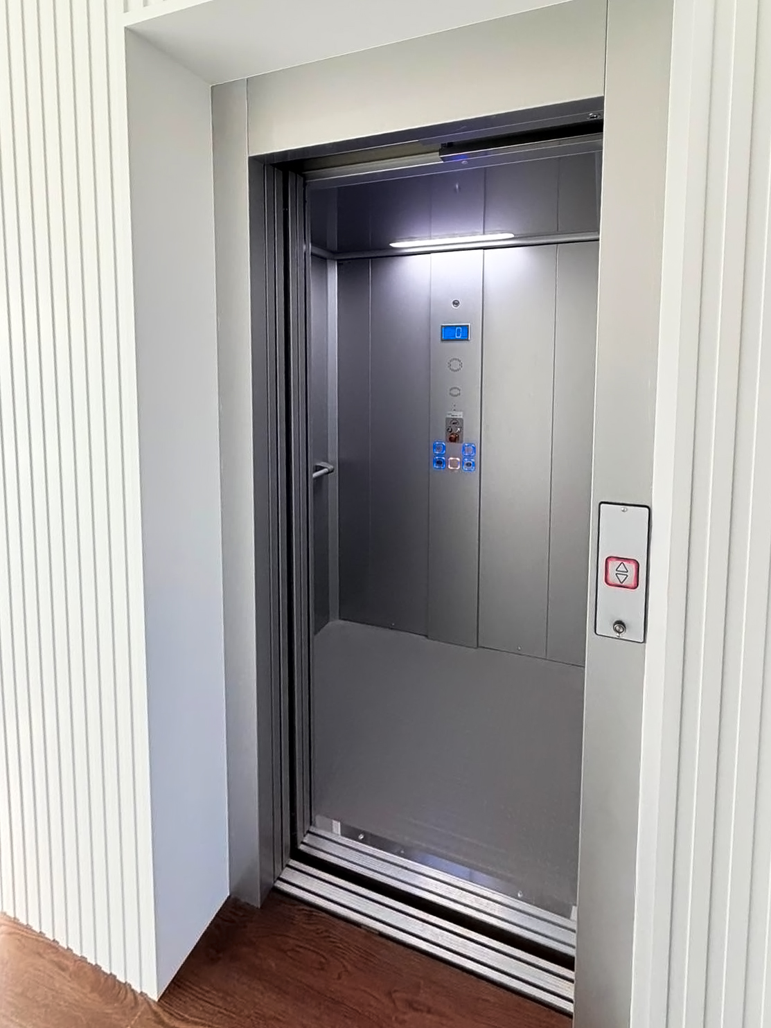 Open modern home elevator with stainless steel interior and wooden floor outside.