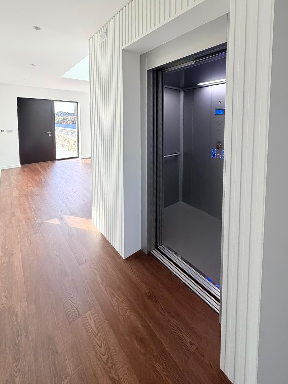 Modern home interior with an open elevator featuring stainless steel walls and a wooden floor leading to a black entrance door.