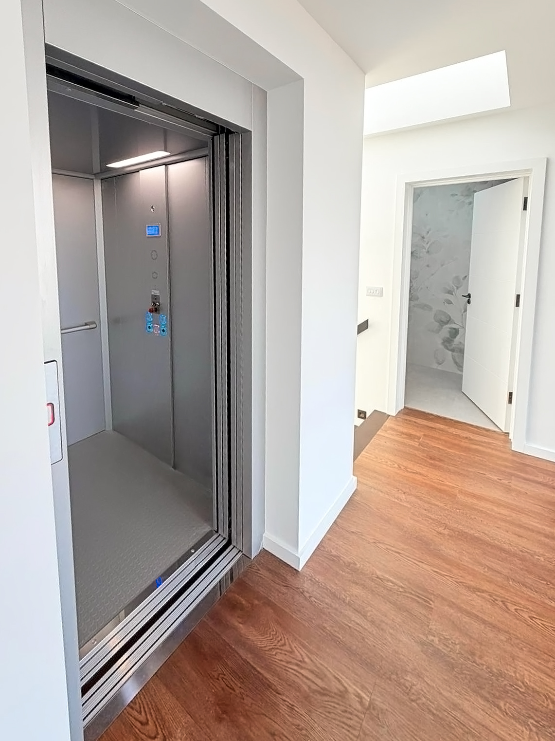 Modern home elevator with open door in a hallway with wooden flooring and a partially open white door leading to a room with floral wallpaper.