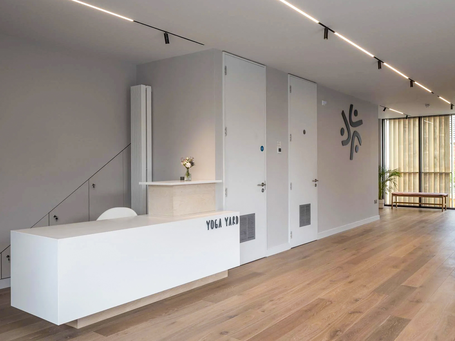 Modern yoga studio reception area with white desk labeled Yoga Yard, wooden flooring, two closed doors, and a bench near large windows.