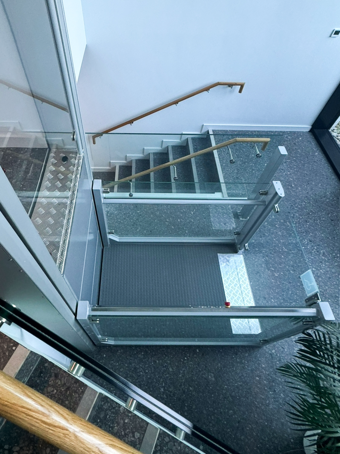 Indoor staircase with glass railing next to a wheelchair lift and large floor-to-ceiling windows showing an outdoor view with plants.