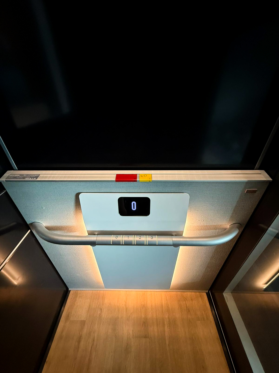 Elevator interior with wooden floor, illuminated handrail marked with floor numbers 0 to 3, and digital display showing floor 0.