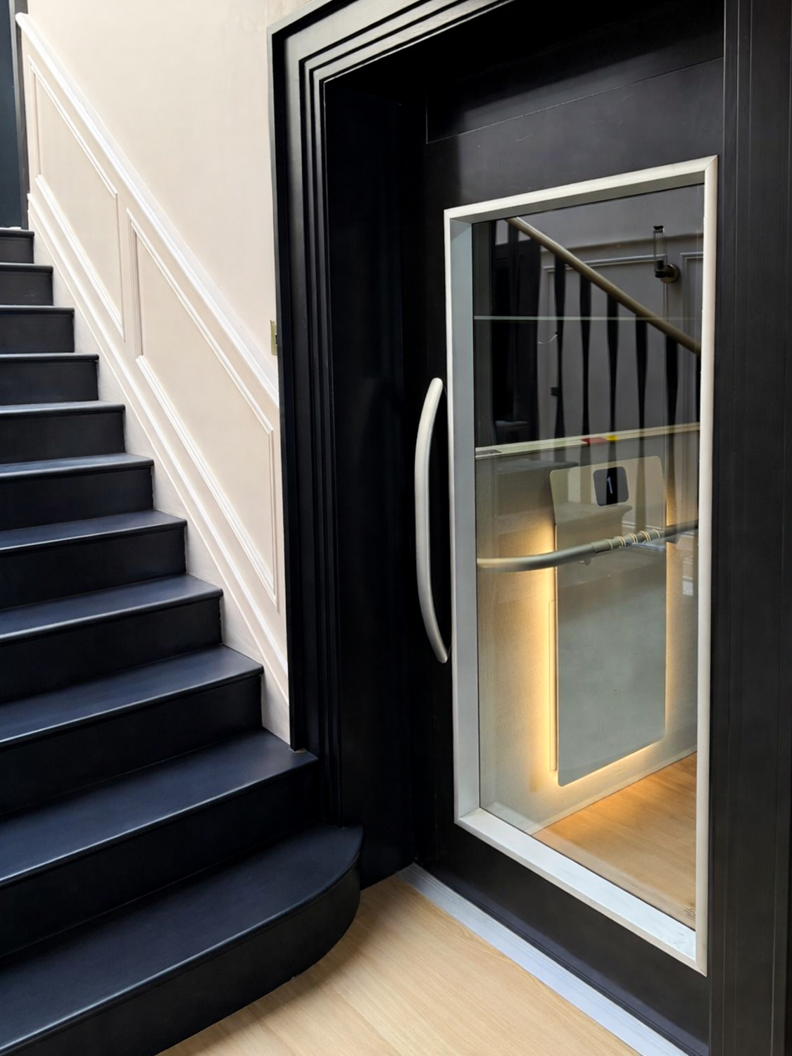 Modern black staircase next to a glass door with a silver handle revealing a small, illuminated platform lift inside.