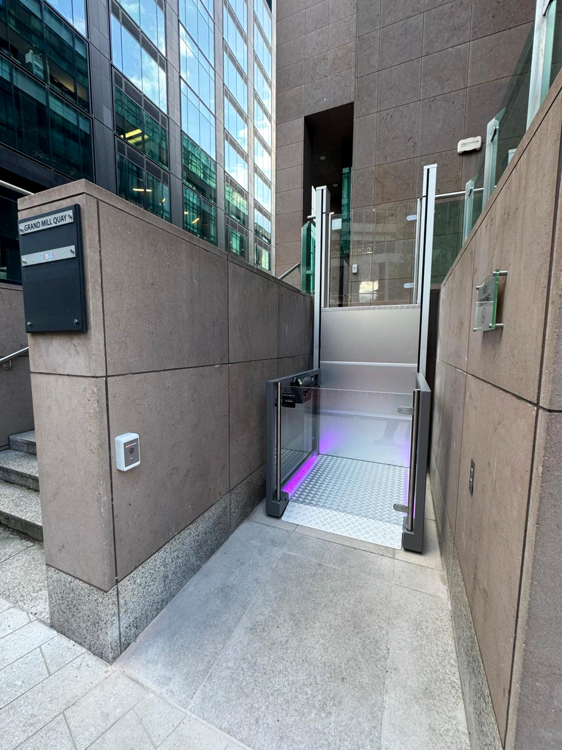 Outdoor wheelchair lift with glass panels and purple lighting, installed between two stone walls near building entrance.