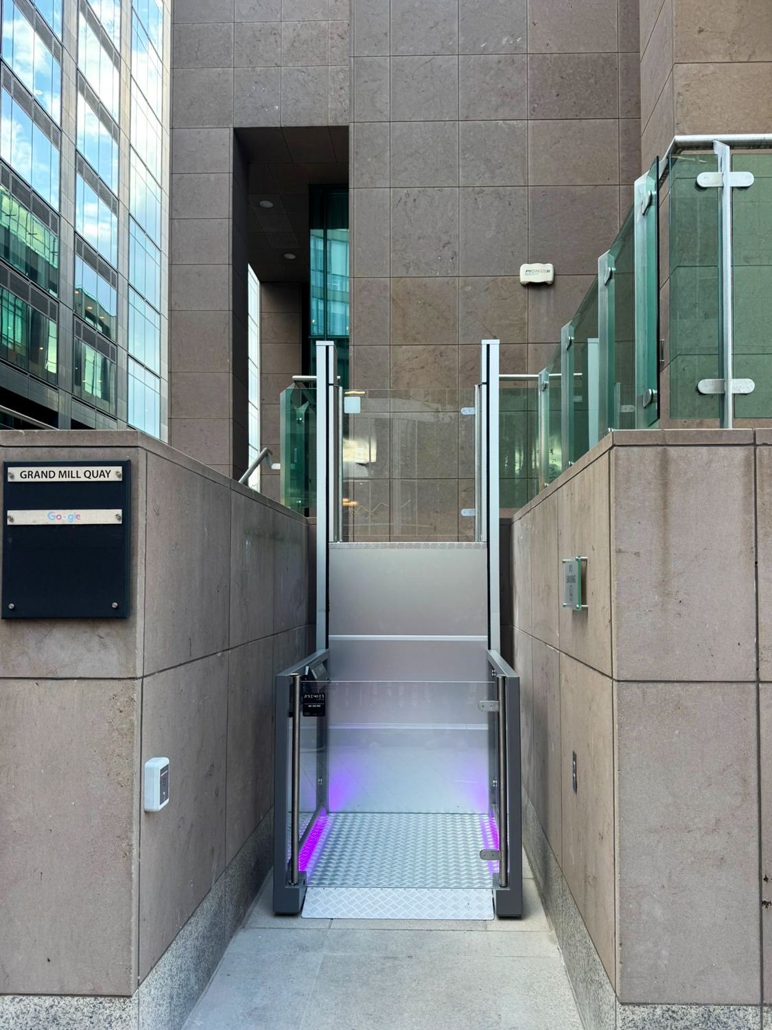 Outdoor wheelchair lift with glass sides and purple lighting, installed between brown stone walls near a building entrance.