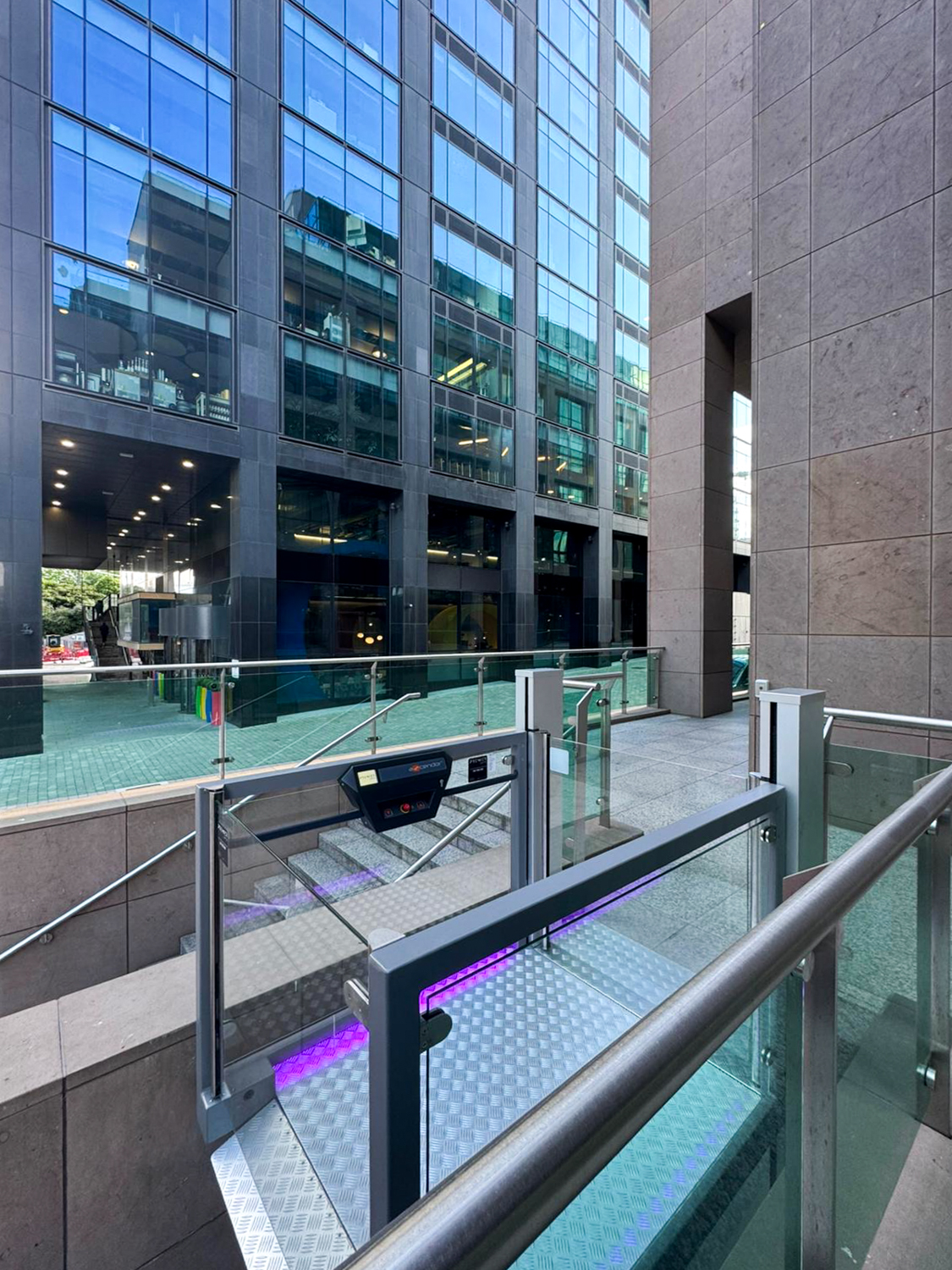 Modern wheelchair lift platform with glass and metal railings next to stairs, outside a building with reflective glass windows.