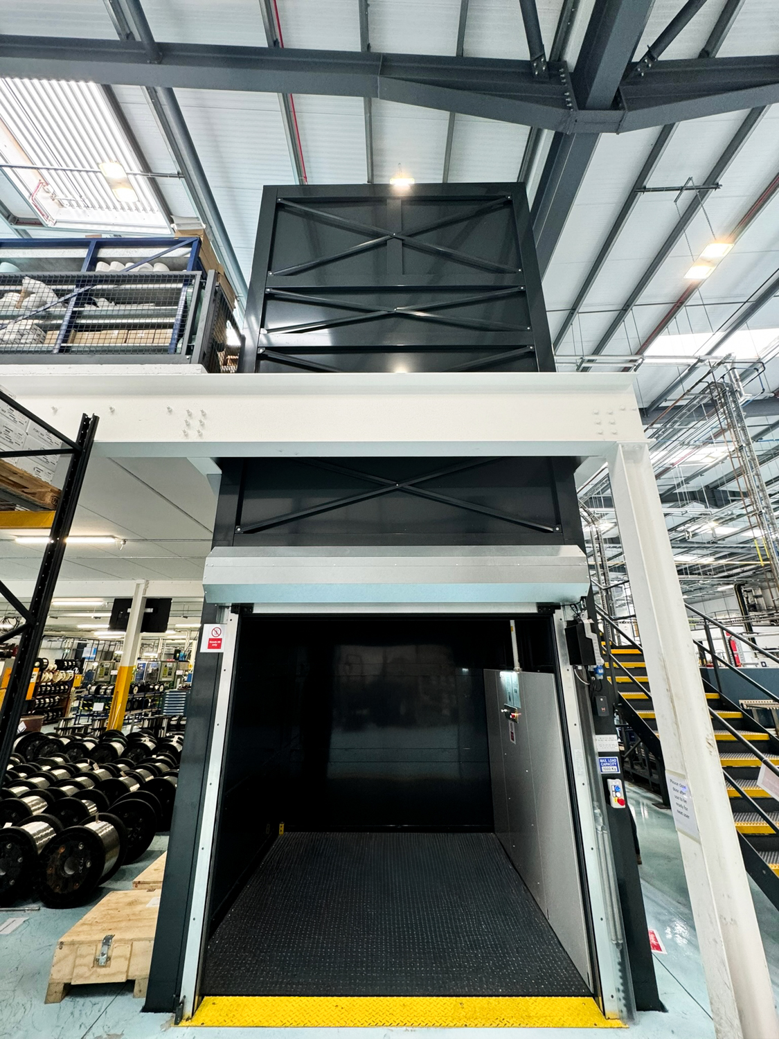 Interior of a metal goods lift with gray panel walls and a control panel with buttons and warning signs.